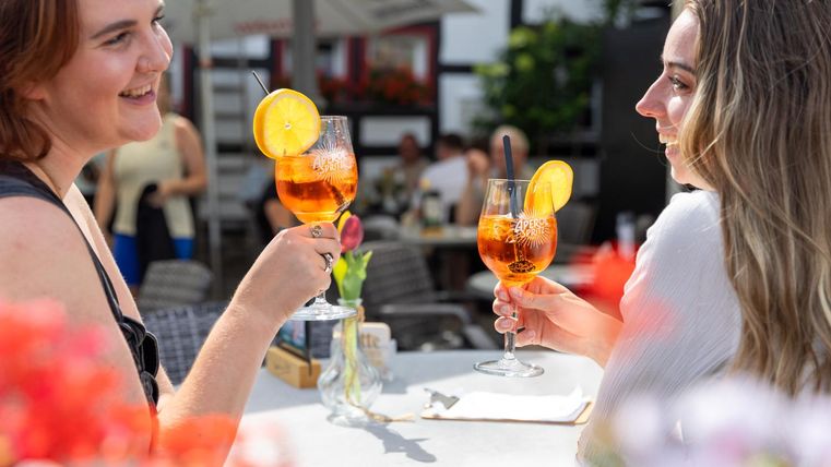Twee vrouwen proosten met kleurrijke dranken. Op de achtergrond zijn mensen en bloeiende planten te zien.