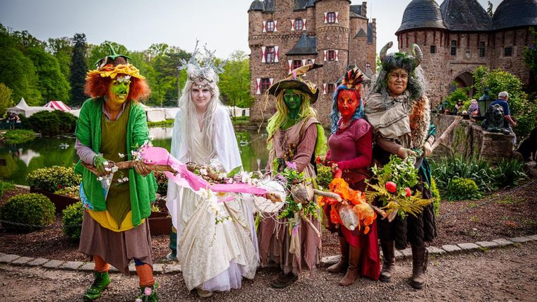 Un groupe de personnes déguisées de manière fantaisiste se tient devant un château.
Elles portent des costumes colorés et tiennent des bâtons décorés, entourés d'un paysage pittoresque.