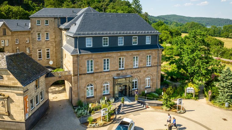 Een historisch gebouw met modern design omgeven door groene landschap. Bezoekers staan voor de ingang en een auto parkeert op het terrein.