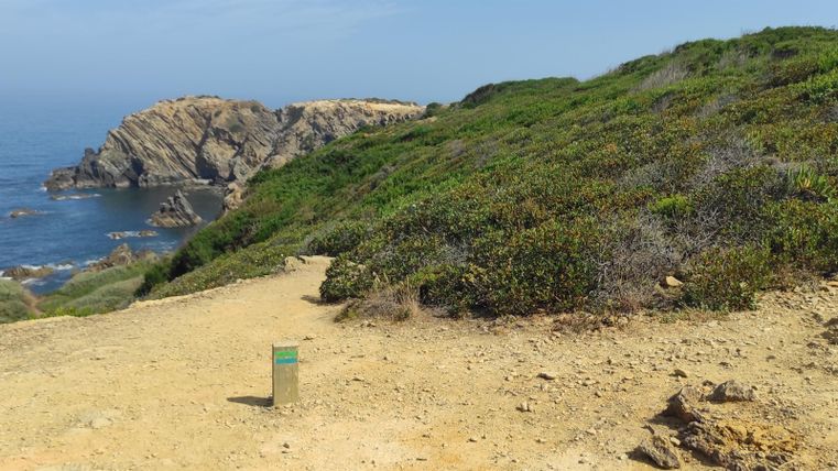 Ein Weg führt zu einer Küste mit felsigen Klippen. Im Hintergrund ist das blaue Meer und bewachsene Hügel zu sehen.