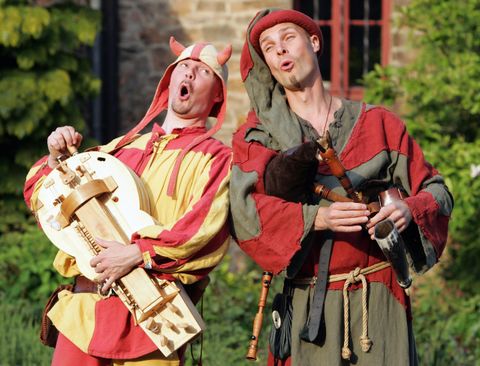 Deux artistes vêtus de costumes médiévaux chantent et jouent des instruments de musique. Ils affichent des expressions joyeuses et se tiennent devant un décor verdoyant.