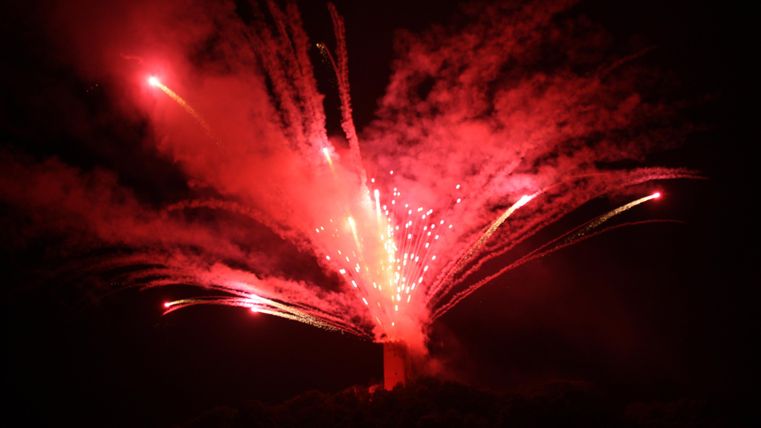 A spectacular fireworks display with bright red and white sparks lights up the night sky. The explosions create an impressive visual spectacle over the landscape.