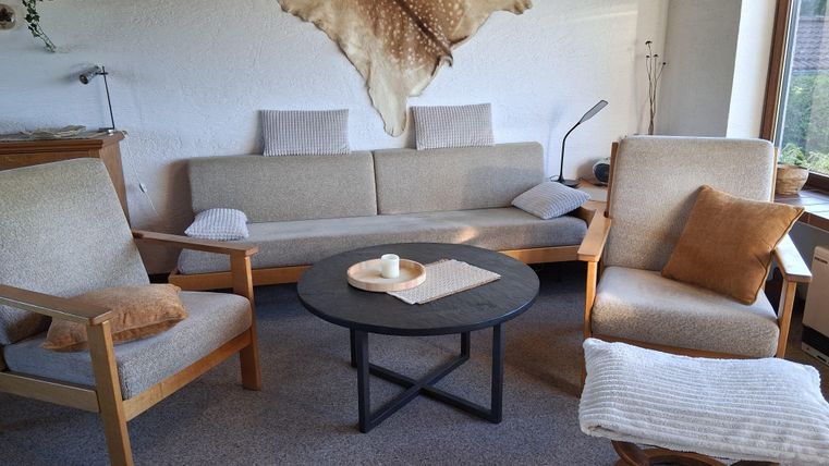 Een gezellige woonkamer met een beige bank en twee stoelen. Een ronde tafel staat in het midden, en een decoratieve wanddecoratie is zichtbaar.