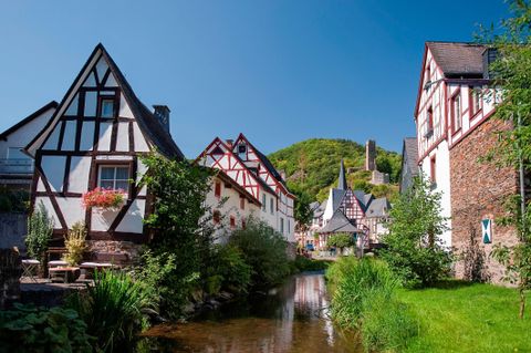 A picturesque village with half-timbered houses by the bank of a clear stream. In the background, a wooded hill can be seen.