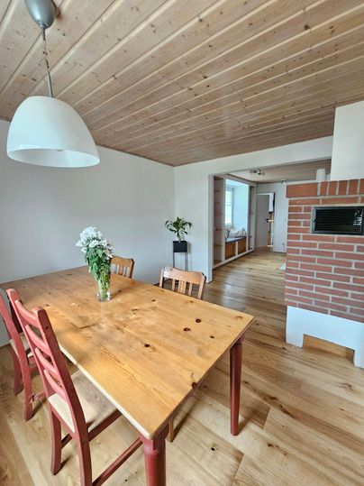 Een lichte eetkamer met een houten tafel en rode stoelen. Op de achtergrond zie je planten en een open haard.