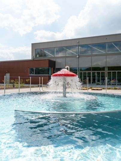 Une piscine moderne avec un toit en spray rouge qui éclabousse de l'eau. À l'arrière-plan, un bâtiment avec de grandes fenêtres est visible.