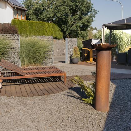 Eine moderne Gartenlandschaft mit einer Holzliege auf einem runden Holzdeck. Im Hintergrund sind hohe Gräser und ein Pavillon zu sehen.