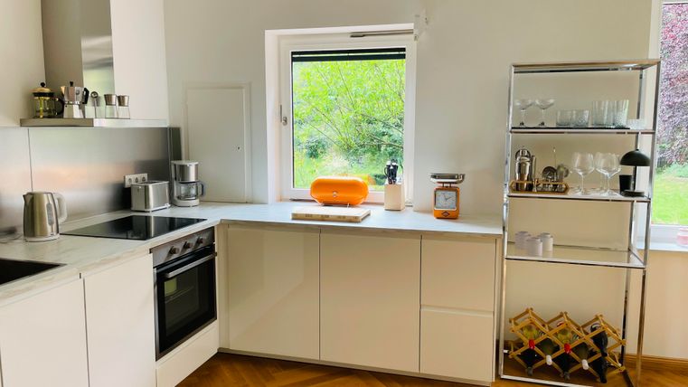 Une cuisine moderne avec des meubles blancs et un élégant parquet en bois. Sur le plan de travail, il y a une serviette orange, tandis que la fenêtre offre une vue sur le jardin.