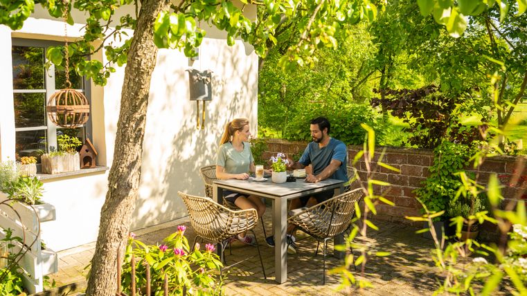 Twee mensen zitten aan een tafel in de tuin, omringd door groene planten en bomen.