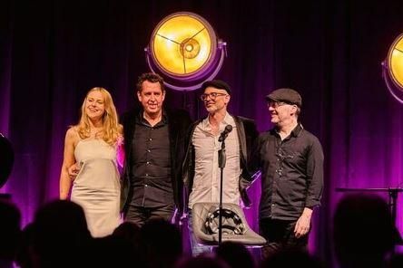 A group of four people stands on a stage in front of a purple-lit background. They appear relaxed and are smiling at the camera.