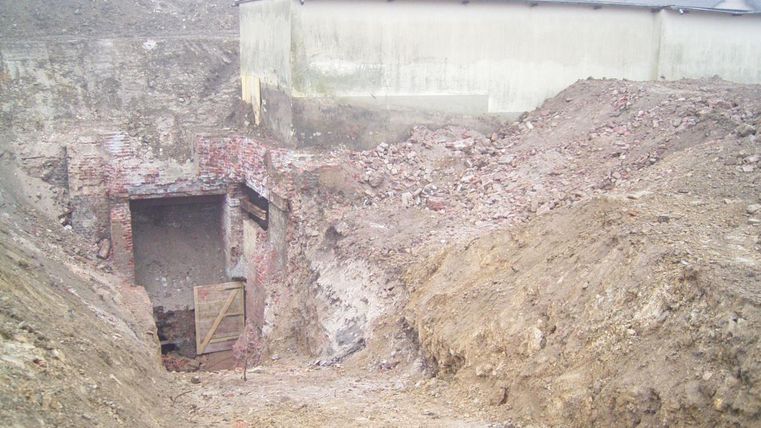 A construction site with a deep, empty trench and broken bricks. In the background, a gray wall of a building is visible.