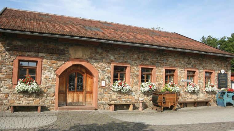 <p>Ein eingeschossiges längliches Gebäude aus Bruchsteinen und mit einem rotem Ziegeldach. Der linksgelegene Eingang hat einen Rundbogen aus rötlichem Sandstein.Die Holztür ist im oberen Bereich aus Glas und mit einem Schmiedegitter verziert. Rechts und links sind 2 feststehene Holzelemente. Links des Einganges gibt es 1 Fenster, rechts davon fünf, vierreckige Sprossenfenster, die ebenfalls von Sandsteinen umrahmt sind. Vor den Fenstern sind Blumenkästen mit weißen und roten Blumen. Der Hof vor dem Gebäude ist gepflastert.</p>