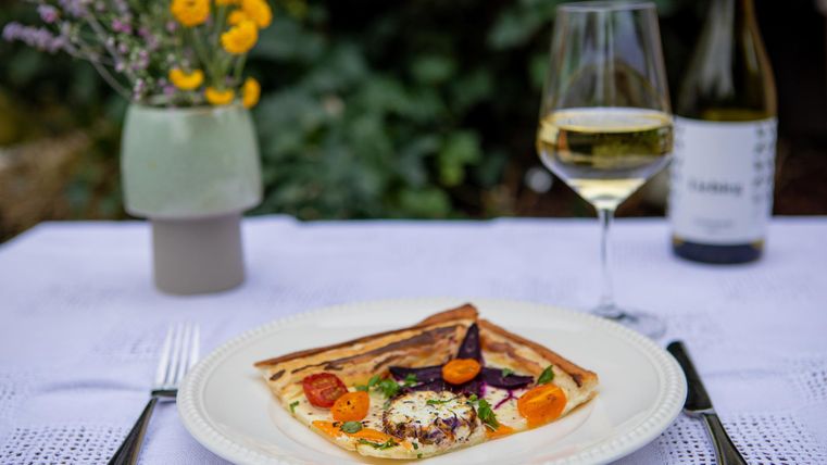 Een bord met een kleurrijke bladerdeegkruidenpasta, versierd met kleurrijke groenten en kaas. Naast staat een glas witte wijn en een vaas met verse bloemen.