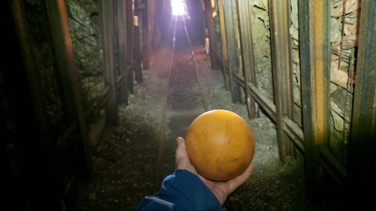 Eine Person hält eine orangefarbene Kugel in einem alten Tunnel. Am Ende des Tunnels ist helles Licht sichtbar.