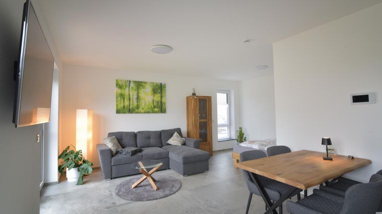 Ein modernes Wohnzimmer mit einer grauen Couch und einem Holztisch. Eine Pflanze und ein Bild mit Waldmotiven sorgen für eine gemütliche Atmosphäre.