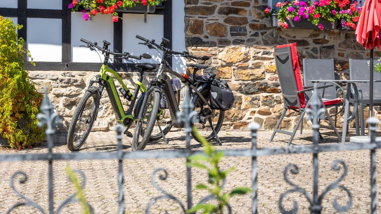 Two bicycles are standing by a stone wall next to a cozy outdoor area. Colorful flower boxes and umbrellas give the scene an inviting atmosphere.