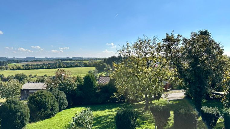 Un paysage vaste avec des prairies vertes et des arbres sous un ciel bleu clair. À l'arrière-plan, on peut voir des collines douces et quelques bâtiments.
