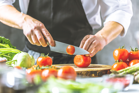 A cook is slicing tomatoes on a wooden board. Fresh vegetables and herbs are lying around him.