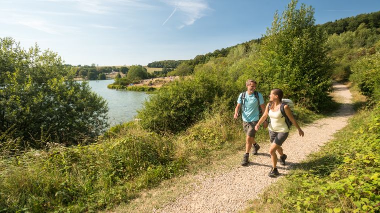 Zwei Wanderer auf dem Weg neben einem See rechts udn links vom Weg grüne  Sträucher , heller Himmel