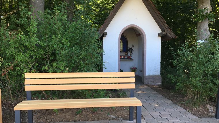 Eine kleine Kapelle mit Bank im Vordergrund, umgeben von Bäumen.