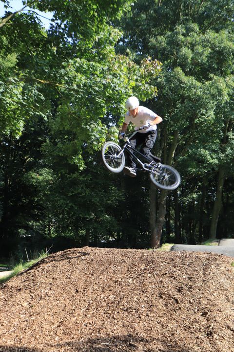 Un cycliste BMX saute par-dessus un tremplin sur une colline de skatepark. À l'arrière-plan, on peut voir des arbres et un environnement verdoyant.
