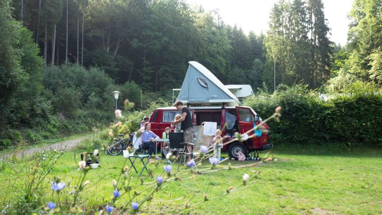 Une scène de camping confortable avec un camping-car rouge et des gens qui se détendent à l'extérieur. Les environs sont verts avec des arbres en arrière-plan.