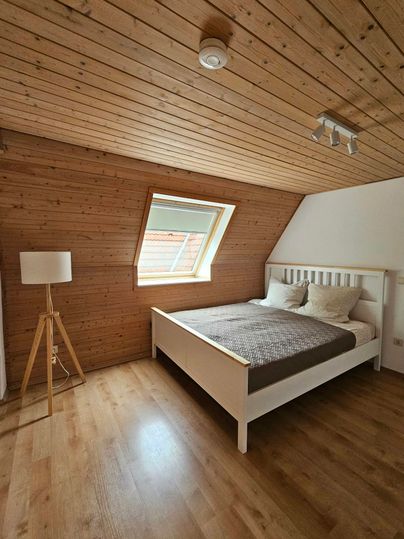 Een gezellig slaapkamer met een witte bed en een moderne lamp. De muren zijn van hout en er is een dakraam dat daglicht binnenlaat.