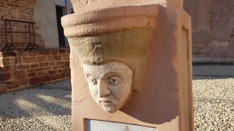 Een kunstzinnige sculptuur met een gezicht en een hoed van steen. De figuur staat tegen een muur in een historisch rijke omgeving.
