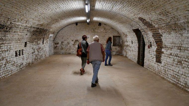 Une pièce vide avec un haut plafond voûté et des murs en briques blanches. Trois personnes traversent la pièce.
