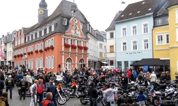 Eine lebhafte Stadtplatz mit vielen Motorrädern und einer großen Menschenmenge. Im Hintergrund sind historische Gebäude in verschiedenen Farben zu sehen.