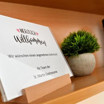 Ein Willkommensschild auf einem Ablagetisch. Darunter stehen freundliche Grüße vom Team der St. Martin Gastronomie.