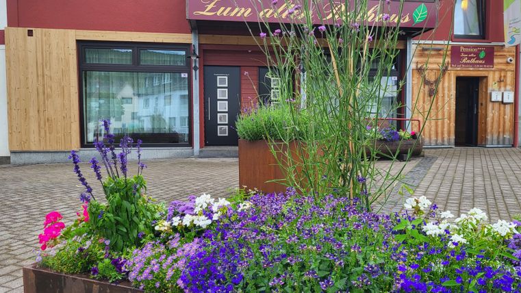 A blossoming flower pot, filled with colorful flowers, stands in front of a building. The sign "Zum alten Posthaus" is visible above the entrance.