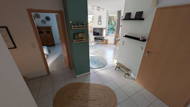 Een moderne gang met lichte muren en een tegelsvloer. Op de achtergrond is een gezellige woonkamer te zien met planten en een televisie.