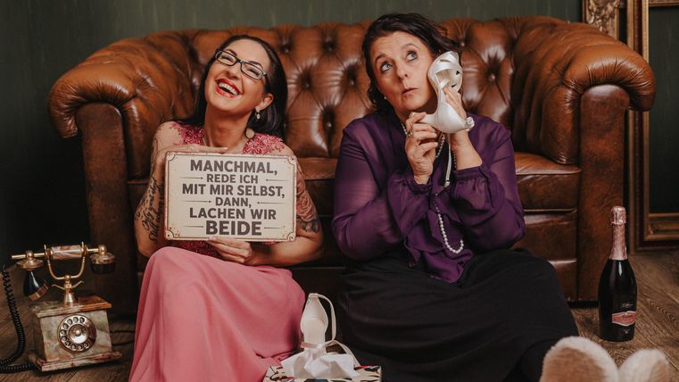 Two women are sitting, laughing and thoughtfully, on a leather sofa.