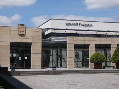 Ein modernes Gebäude mit Glasfassade und einem Eintragungszelt. Vor dem Gebäude stehen gepflegte Pflanzen.