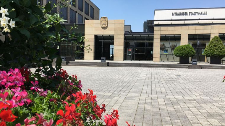 Ein modernes Gebäude mit Glasfassade und einem Eingang. Im Vordergrund blühen bunte Blumen auf einem Platz.