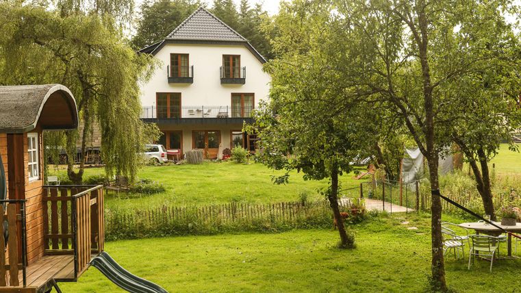 Ein gemütliches Haus umgeben von grünem Rasen und Bäumen. Im Vordergrund befindet sich eine Holzterrasse mit einer Hängematte.