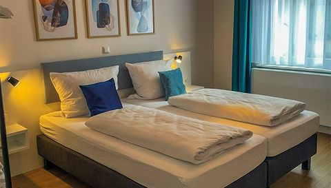 Een gezellige slaapkamer met twee bedden en lichte kussens. Aan de muur hangen decoratieve schilderijen, en het natuurlijke licht stroomt door het raam.