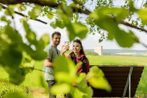 Two people take a selfie in front of the Bruder Klaus Chapel, surrounded by green leaves.
