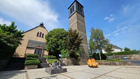 Un bâtiment historique doté d'une haute tour et entouré d'une végétation luxuriante. La place située devant le bâtiment est pavée et ornée d'une petite sculpture.