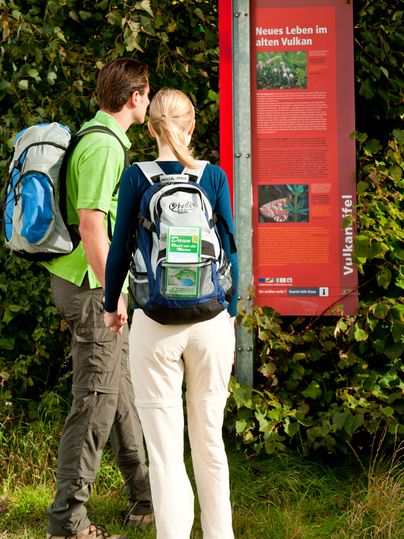 Zwei Wanderer lesen ein Informationsschild im Grünen.