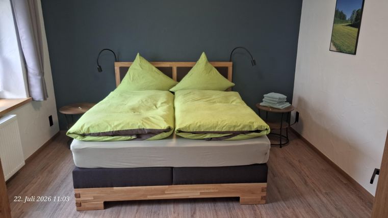 A cozy bedroom with a large, comfortable bed and green pillows. Next to the bed is a small nightstand with stacked towels.