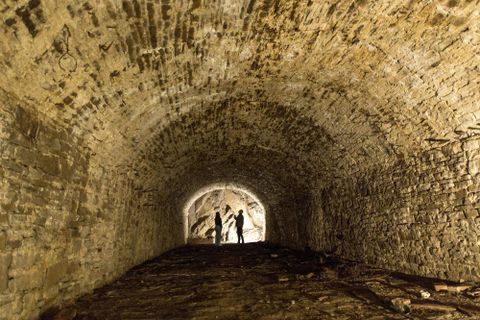 Ein dunkler Tunnel mit steinernen Wänden und einem gewölbten Dach. Zwei Silhouetten stehen am Ende des Tunnels und scheinen sich zu unterhalten.