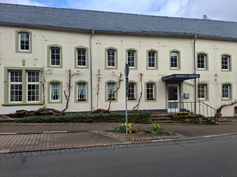 The image shows the facade of a hotel with several windows and an entrance. Plants are growing around the building, and in the foreground, flowers can be seen.