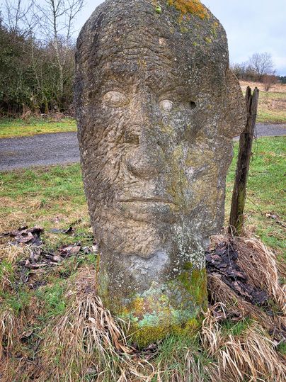 Eine große Steinskulptur in Form eines Gesichts steht auf einer Wiese. Im Hintergrund ist eine bewaldete Landschaft und ein grauer Himmel zu sehen.
