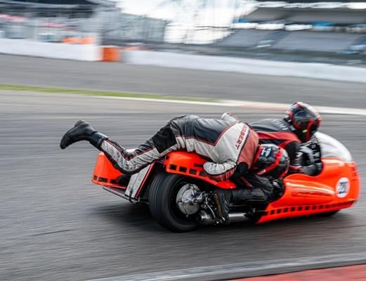 Een motorfietscombinatie op het circuit met een coureur die zich aerodynamisch op het voertuig legt. De scène laat snelheid en dynamiek zien.