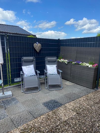 A cozy terrace with two loungers and blooming plants in a flower pot. The sky is blue with some clouds.