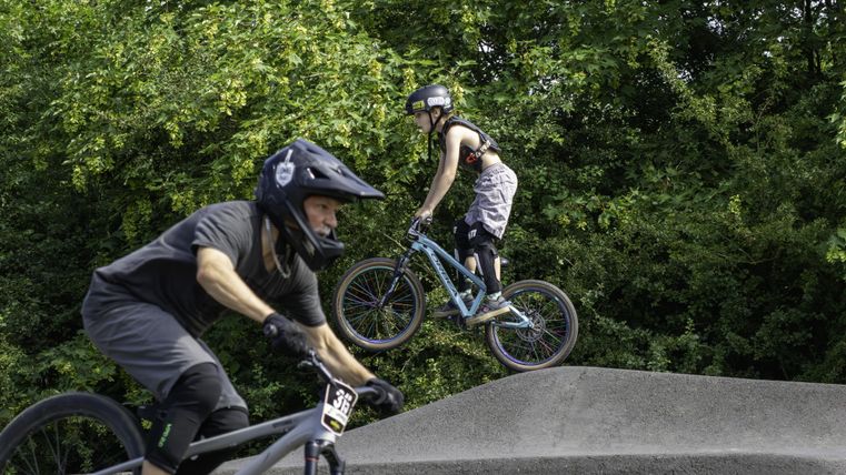 Deux cyclistes exécutent des tricks acrobatiques sur un sentier. L'environnement est vert et il fait un temps ensoleillé.