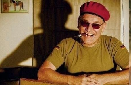 Ein lächelnder Mann mit Sonnenbrille und rotem Barett sitzt an einem Tisch. Er hält eine Zigarette im Mund und scheint entspannt.