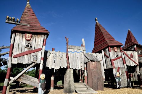 Ein Spielplatz mit Holzburgen und roten Dächern. Der Himmel ist blau und die Umgebung ist sonnig.
